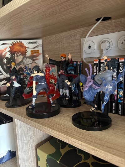 This image captures a display of collectible anime figurines and media on a light brown, wood-grain laminate shelf in Besançon, France. A total of four primary figures are arranged on individual black circular bases. From left to right, a figure resembling Dabi from My Hero Academia is posed with a blue flame effect emanating from his left hand, next to a figure resembling Itachi Uchiha from Naruto, partially obscured by a red flame-like construct. Further right, a figure resembling Pain (Nagato) from Naruto stands with arms slightly elevated. On the far right, a figure resembling Sasuke Uchiha from Naruto is depicted in mid-motion, with a blue electrical effect emanating from his right hand.
In the background, behind the figures, a large "Bleach" DVD/Blu-ray case featuring Ichigo Kurosaki is visible on the left. To the right, a row of manga volumes, including titles such as "Jujutsu Kaisen" by Gege Akutami and "My Hero Academia," are stacked vertically. Above the manga volumes, a white triple-gang wall outlet plate is visible, featuring three French Type E power sockets and a coaxial TV port. A white power cable extends from a circular cutout in the shelf above and plugs into one of the power sockets. The shelf unit continues below the depicted level, revealing a portion of another shelf and a green and black camouflage-patterned box.