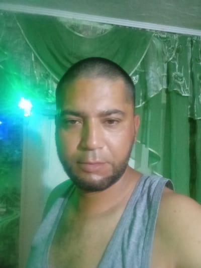 A close-up, waist-up selfie shows a man with short hair and a beard, wearing a grey tank top. He is standing in front of green curtains. On the left side of the image, there is a bright green light source with some blue accents, possibly from string lights or a projector. The background curtains have a floral pattern. The man is looking directly at the camera. There is no discernible indication of the city of Edineț, Moldova within the image itself.