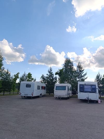 Three white travel trailers are parked on an asphalt lot under a bright blue sky with scattered white clouds. The setting appears to be an outdoor parking area or campsite, bordered by a line of green trees, including pine and deciduous varieties, and a metal fence in the background on the left.
The left-most caravan is a Bailey Unicorn model, with "UNICORN" and "BAILEY" visible on its rear. Its license plate, "YX 2209 77", indicates a Russian registration (77 is a Moscow region code). The caravan in the center is also white and appears to be an older model. The right-most caravan is a Lunar model, with "LUNAR" visible on its front. All three caravans are unhitched and appear to be supported by leveling jacks. A power cable is visible extending from the right-most caravan.
No people are visible, and no specific activity is taking place other than the caravans being parked. The time of day is daytime, with clear, sunny weather. The overall impression is one of a quiet, possibly recreational, parking area in Ivanovskoe, Russia.