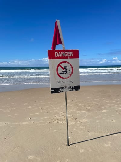 A "DANGER NO SWIMMING" sign with a red and white flag is posted on a sandy beach in the city of Gold Coast, Australia. The sign is weathered and shows some wear and tear. A red circle with a diagonal line across a silhouette of a swimmer indicates the prohibition. The ocean with moderate waves is visible in the background, and the sky is blue with scattered clouds. Footprints are present in the sand, indicating previous activity. The sign is mounted on a metal pole that casts a shadow on the sand.