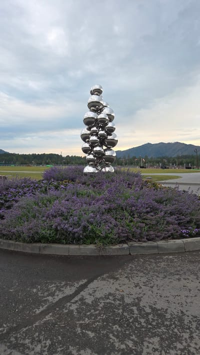 A vertical outdoor shot depicts a striking modern sculpture composed of numerous polished silver spheres connected by dark metal chains, set against a vast, cloudy sky. The sculpture is positioned to the left of the frame, its reflective surfaces mirroring the sky and surrounding elements. To the right, a dense border of vibrant purple flowers, likely lavender or catmint, runs along a paved asphalt path. The path is bordered by a concrete curb. In the background, beyond the flowers and a grassy field, are distant, hazy mountains and trees, suggesting a natural, potentially rural or park-like setting. The lighting suggests it is either early morning or late evening, with soft, diffused light. The sky is filled with various shades of grey and white clouds, with a hint of warm yellow light on the horizon, indicating either sunrise or sunset. There is no visible text in the image. The overall mood is serene and contemplative, highlighting the juxtaposition of modern art with natural beauty.