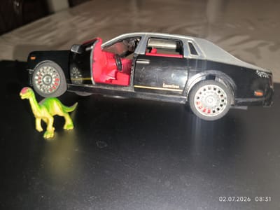 A black toy sedan with a silver roof and red interior sits on a dark, reflective surface. The driver's side door of the car is open, revealing the red interior with a black steering wheel. A small, green toy dinosaur with a pink head stands to the left of the car, facing the car. The background is blurred and shows a textured surface. A timestamp in the bottom right corner reads "02.07.2026 08:31". The scene is indoors and appears to be a tabletop. There is no indication of the city of Kattasu, Uzbekistan in the image.