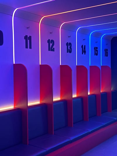 The image shows a tiered seating arrangement in a bowling alley located in Dartford, United Kingdom. The seating platforms are numbered from 11 to 16, each with a corresponding number painted in black on a white surface. Red dividers separate each platform, and blue padded seats are situated on each level. LED lights line the edges of the tiers, transitioning from red and orange hues on the lower numbers to blue on the higher numbers. Each platform has a small hook at the front. The general lighting casts a blue and purple tone over the entire scene.