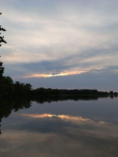 A serene lake reflects the soft, diffused light of a cloudy sunset. The sky, predominantly light blue and grey with wispy clouds, is bisected by a horizontal band of warmer tones where the sun is peeking through. The treeline along the far bank is dark and silhouetted against the lighter sky, with a few taller trees visible on the left. The water is calm, creating a mirror image of the sky and the distant shore, with the reflection of the sunset's warm light adding a golden hue to the water's surface. The overall impression is one of tranquility and natural beauty at the end of the day.