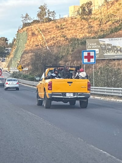 A yellow pickup truck is on the road, filled with people in the back. The truck has a license plate. A red cross sign is visible on the side of the road, along with a real estate advertisement. Several vehicles are visible on the road, including a white car and several others in the distance. The landscape includes a hillside with trees and vegetation.
