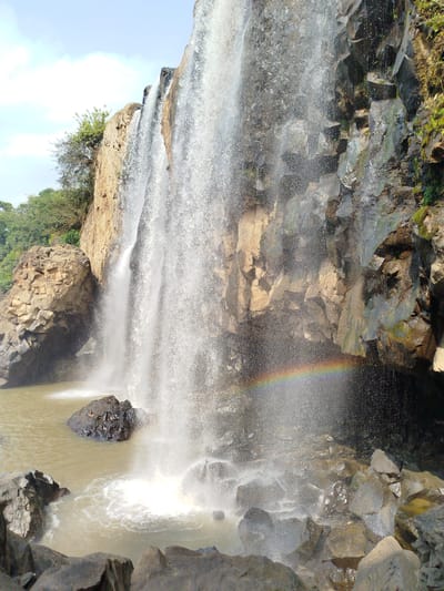 The image shows a waterfall cascading over a rocky cliff into a pool of muddy water. A small rainbow is visible in the mist created by the falls. The surrounding environment consists of trees and vegetation on the cliff edges, and rocks in the foreground. The location is presumably in or near the city of Đà Giang, Vietnam, but no specific landmarks are visible to confirm the exact location.