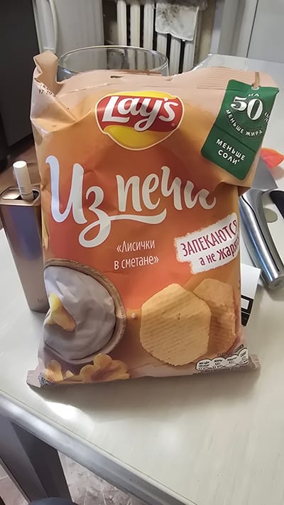 The image shows a bag of Lay's chips with the flavor "Chanterelles in sour cream" on a white table. The bag is orange and has the Lay's logo in red and yellow. There is a green tag attached to the bag that says "50% less fat" and "less salt". Next to the bag is a silver device that appears to be an e-cigarette or vaporizer. In the background, there is a glass and a radiator. The lighting in the scene is bright and appears to be indoors. The image does not contain any explicit information about its location in Yuryev-Polsky, Russia.