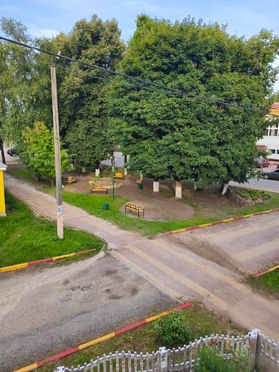 This media file captures an outdoor scene in Spas-Demensk, Russia, likely on a partly cloudy day with soft lighting suggesting either early morning or late afternoon. The perspective is from above, looking down onto a paved area and a small park-like space.

The main subjects are natural elements and urban infrastructure. Large, leafy green trees dominate the background, casting shadows on the ground. In the foreground, a yellow and red striped curb runs along the edge of a gravelly path and a pedestrian crossing made of light-colored interlocking pavers. Scattered throughout the park area are several empty wooden benches, some surrounding a small circular stone structure. A concrete utility pole with wires stretches across the frame, with a small white object, possibly a bird, perched on one of the wires. In the distance, the edge of a building with a yellow facade is visible, along with a street where a few cars are parked or driving. A white picket fence with some flowering plants is in the bottom right corner.

There is no discernable activity or event taking place; the scene appears peaceful and unoccupied. The overall impression is one of a quiet, possibly residential or public area, integrated with nature. No visible text is present in the image.