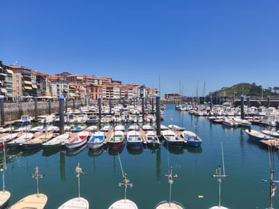 A wide shot captures the picturesque harbor of Lekeitio, Spain, under a clear blue sky. The foreground is dominated by the tranquil, deep blue water of the bay, reflecting the surrounding scene. Several small to medium-sized boats, predominantly white with colorful accents, are docked in neat rows. Some boats are equipped with masts, indicating they are sailboats, while others appear to be motorboats. Small weather instruments are visible on poles in the water in the foreground.
Behind the boats, a long, stone quay stretches across the mid-ground, lined with dark vertical posts that serve as mooring points. Along the quay, a promenade is visible with glimpses of outdoor seating areas and people strolling.
The background is defined by a vibrant line of multi-story buildings that form the charming townscape of Lekeitio. These buildings showcase a variety of architectural styles, with colorful facades ranging from warm terracotta to pastel hues, and many feature terracotta-tiled roofs. Yellow and red awnings are visible over windows and balconies.
Further in the distance, a verdant, hilly landscape rises, topped with trees, and a bridge or elevated walkway is visible in the distance. The overall impression is one of a serene and bustling coastal town on a bright, sunny day.