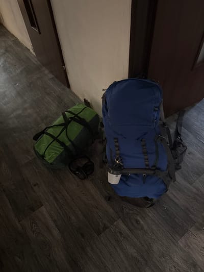 The image shows two backpacks and a pair of headphones on a dark wood-grain floor. The larger backpack, in the upper portion of the image, is blue and appears to be a hiking or camping backpack with multiple straps and buckles. A white cylindrical object, possibly a water bottle or flashlight, is attached to its side. Below the blue backpack, a green bag is visible, secured with black straps. To the right of the green bag, a pair of black headphones rests on the floor. The scene is indoors, with a wall and what appears to be a dark-colored door or furniture in the background on the left. The lighting suggests it is likely daytime, but the specific time of day or weather conditions cannot be determined. The overall impression is one of preparation for travel or an outdoor activity. No visible text is present in the image.