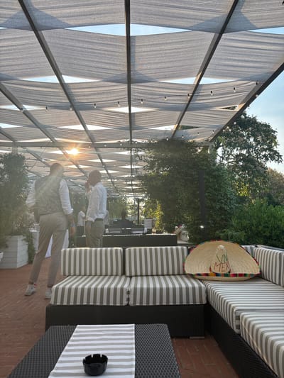 Two men are seen in an outdoor seating area, possibly a restaurant or cafe, with a shaded canopy overhead. The canopy is made of light-colored fabric stretched over a metal frame, with string lights illuminating the space. To the right, there is an outdoor sofa with striped cushions, and on top of the sofa sits a sombrero. The ground is paved with bricks. The sun is low in the sky, casting long shadows and creating a warm, golden light, suggesting it is late afternoon or early evening. The foliage of trees is visible in the background. One man, wearing a white shirt and khaki pants, is standing and looking towards the other man. The second man, dressed in a vest over a white shirt and khaki pants, is also standing and appears to be gesturing. The overall atmosphere seems relaxed and social. The sombrero has colorful embroidery and the word "MEX" is visible.