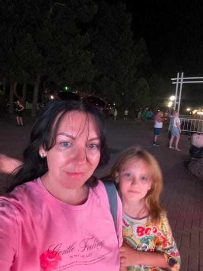 In the foreground of the image, a woman with long dark hair is taking a selfie. She is wearing a pink t-shirt with "Gentle Feelings" printed on it. To her right, a young girl with blonde hair and blue eyes is visible, wearing a colorful t-shirt with the word "Boo" printed on it. They appear to be outdoors at night, as evidenced by the dark background with trees and streetlights. In the background, several other people are walking along a paved pathway. The setting is the embankment of the city of Krinitsa, Russia.
