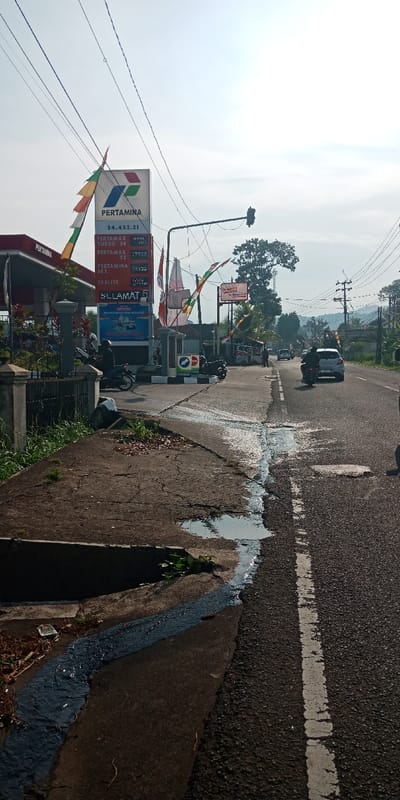A daytime street view in Cijedil, Indonesia, captures a Pertamina gas station and an active roadway under a hazy sky.
In the foreground, a cracked concrete sidewalk with sparse vegetation includes a shallow drainage channel carrying a dark liquid that spills onto the asphalt road, forming a distinct dark streak. Small puddles are present on the sidewalk and at the edge of the road. A black metal fence and concrete pillars are visible on the far left.
The midground features the entrance to a Pertamina gas station on the left. A tall, illuminated Pertamina sign displays fuel prices for Pertamax Turbo 98, Pertamax 92, Pertamina Dex, and Dexlite, along with the station number 34.432.21. Colorful striped banners are affixed to the sign and draped across the station entrance. A red and white Pertamina canopy is partially visible. Two motorcycles are parked or stationary near the pumps, with at least two individuals present, one mounted on a motorcycle. Overhead, a black traffic light hangs above the road. Further down the street on the left, signs for Indomaret and Alfamart are visible.
The asphalt road, marked by a faded white line, extends into the background. A black motorcycle with a helmeted rider and a light-colored car are observed moving away from the viewer on the right side of the road. Additional vehicles are visible in the distance. The background consists of a treeline, utility poles with numerous overhead power lines, and distant hills under a light, hazy sky.