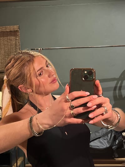 A fair-skinned person is taking a selfie with a dark green smartphone. The person has shoulder-length blonde hair, pink lips, and several rings and bracelets on their hands and wrists. They are wearing a black sleeveless top. The background includes a muted blue wall, a decorative woven item, a silver pole, and the person's gray backpack and frame. The location is not identifiable as Livingstone, Zambia.
