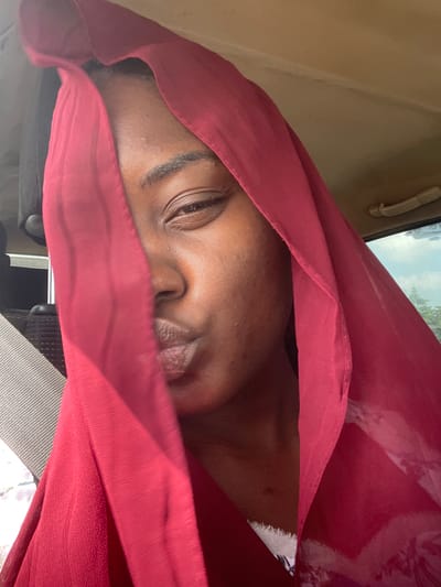 A close-up, slightly out-of-focus shot depicts a person with brown skin partially veiled by a red fabric, likely inside a vehicle, as evidenced by the vehicle's interior features in the background. The person's left eye is visible and open. The nose and mouth are partially covered by the fabric. The background reveals an interior grab handle and a glimpse of the outside through the car window. There is not enough detail to place the location in the city of Dokwa, Nigeria.