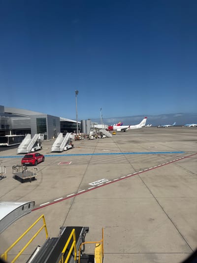 The image depicts an active airport apron scene under a clear blue sky, situated at Tenerife South Airport (TFS) in Granadilla de Abona, Spain.
In the immediate foreground, the perspective is from an aircraft or jet bridge, showing a portion of a white aircraft wing on the lower left and a yellow and black baggage conveyor belt with adjacent stairs on the lower right.
The midground features a white Norwegian Air Shuttle aircraft (likely a Boeing 737) with a distinctive red and blue tail livery (including the word "Norwegian" and a portrait figure), parked at a gate and connected to a jet bridge. Behind this aircraft's tail, a portion of another aircraft with a red and yellow tail, characteristic of Southwest Airlines, is visible. Further in the distance on the tarmac, two additional light blue aircraft are visible.
Numerous pieces of ground support equipment are scattered across the concrete apron, including two prominent mobile passenger stair units (white with metal steps), several smaller tugs or tractors, and baggage carts. A red Mercedes-Benz car, branded with "etnet.com" on its side, is parked on the apron near the mobile stairs.
To the left, a modern airport terminal building with grey paneling and large glass windows forms the backdrop. Several tall, metal light poles are erected across the apron. The concrete surface is marked with various painted lines: blue striped lines, a prominent red and white dashed line, and the white Spanish word "ESPERA" (meaning "WAIT"). No individuals are clearly discernible in the scene; however, the presence of equipment implies ongoing airport operations such as servicing or passenger transfers.