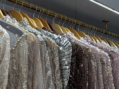 The image shows a row of beaded and sequined garments hanging on wooden hangers from a metal rod, likely in a boutique or a clothing store. The garments appear to be dresses or tops, with intricate embellishments of beads, pearls, and sequins in various shades of silver, gray, and purple. The background is a dark, solid wall, with a strip of white LED lighting visible at the top. The scene is well-lit, highlighting the sparkling details of the clothing. There are no people visible in the image. The location is not specified in the image.
