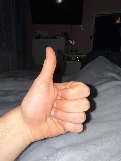 A close-up image shows a human hand, specifically a right hand, giving a thumbs-up gesture. The hand is resting on a soft, gray surface with a plush texture. The background is dark and out of focus, showing glimpses of a television screen, a white shelf unit, and a wall with what appears to be framed pictures or decorative items. No information is provided that indicates the hand is in Musachevo, Bulgaria.