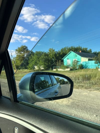 The image captures a view from inside a car, looking out the side window. The scene depicts a rural setting under a clear blue sky with scattered white clouds. A teal-colored house with a black roof and white-framed windows is visible in the background, bordered by a matching teal fence. The house is surrounded by lush green trees and tall, dry grass. A dirt road is visible in the foreground, and a side-view mirror reflects the road behind the car. The interior of the car, including the door panel and window frame, is partially visible. The mirror contains the text "OBJECTS IN MIRROR ARE CLOSER THAN THEY APPEAR." The overall impression is a snapshot of a journey through a quiet, sun-drenched countryside, possibly in the village of Novotolucheyevo, Russia, given the context provided. There are no people or visible actions occurring within the frame.