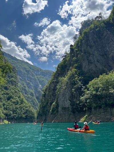 A group of kayakers are paddling on a calm, turquoise lake surrounded by steep, tree-covered mountains. The sky is blue with scattered white clouds. Two kayakers in the foreground are paddling an orange and red tandem kayak. In the middle ground, several other kayaks are visible, some with multiple occupants. The mountains rise dramatically on either side of the lake, with rocky cliffs and dense green foliage. The water is clear and reflects the surrounding scenery.
