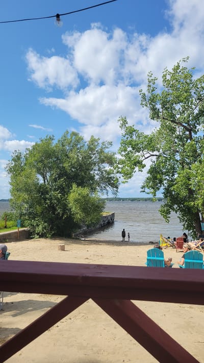 The image depicts a beach scene at the Parc national d'Oka, located in Pointe-des-Cascades, Canada. The foreground is dominated by a maroon-colored railing of a patio or deck, obscuring the lower portion of the sandy beach. Beyond the railing, the beach stretches towards a body of water, likely a lake, with calm, rippling waves.
On the beach, several people are visible. Two individuals, appearing to be an adult and a child, stand near the water's edge, facing the lake. Further back, a group of people are relaxing in beach chairs. Two bright turquoise Adirondack chairs are prominently placed on the sand, each occupied by a person. Other beach chairs, some yellow and some red, are scattered around the area, with more individuals seated in them.
Lush green trees frame the scene, with a large, full tree on the right and another substantial tree on the left, closer to the water. The water extends to the horizon, where a distant treeline is visible under a bright blue sky dotted with fluffy white clouds. A single light bulb hangs from a wire in the upper portion of the frame, suggesting an outdoor lighting fixture. A small, cylindrical tree stump rests on the sand in the middle ground. The overall impression is a relaxed, summer day at a waterfront recreational area.
