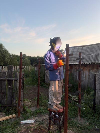 A man wearing a blue shirt, tan pants, a welding mask, and orange gloves stands on a red and black stool welding two rusty metal posts. The welding equipment is connected to a power source visible on the ground. The scene is outdoors, with a wooden fence and a rustic wooden building in the background. Trees and overgrown grass surround the area. The sky is blue with some scattered clouds. This activity appears to be taking place in the rural settlement of Kuseevo, Russia, likely in a backyard or work area.