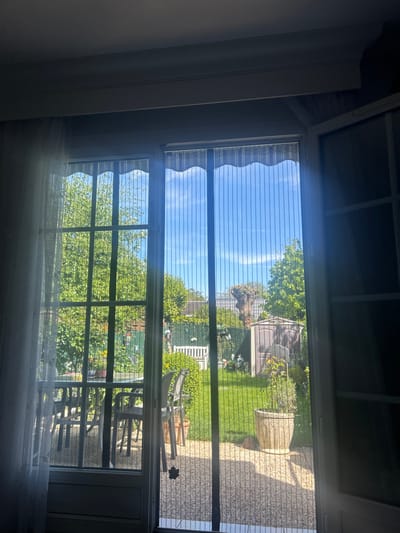 The image shows a view from inside a room looking out through a window, likely in Noisiel, France. The window has a screen and is divided into multiple panes. Outside, there is a garden area visible. The garden features green grass, trees, and plants. Garden furniture, including chairs and possibly a table, can be seen on a paved patio area. In the background, there is a shed-like structure and a fence. The sky is visible through the gaps in the trees, indicating a bright, sunny day. The indoor space appears dimly lit compared to the bright outdoor scene. There are curtains partially visible at the bottom of the window.