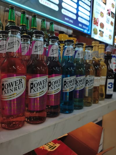 In a retail establishment in Jixi, China, multiple bottles of a beverage labeled "POWER XINRUI Soda Wine" are displayed on a shelf. The bottles are arranged in rows, with varying colors of liquid visible inside, including red, blue, and amber. Some bottles have pink and gold labels with Chinese text, indicating "Classical" and "ALC. 3.0% vol NET.275ml". Behind the display, a digital menu board shows a selection of food items and prices in Chinese. The lighting appears to be artificial, casting reflections on the bottles. The overall scene suggests a point-of-sale environment within a store or restaurant.