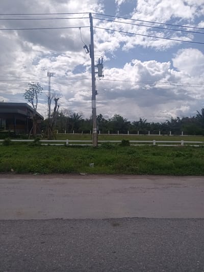 The scene is an outdoor environment in Tha Ruea, Thailand. The foreground is dominated by a two-tone paved road. Beyond the road is a grassy verge, followed by a white fence. Behind the fence is a grassy field with trees. A building is visible to the left, partially obscured by trees.  In the center of the image, a utility pole with electrical hardware stands. Power lines run across the top portion of the image. In the background is a communications tower to the left of the utility pole. The sky is overcast with a mix of white and gray clouds.