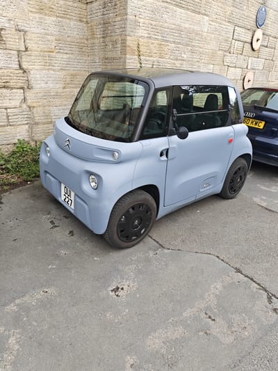 The image depicts a light blue Citroën Ami parked on an asphalt road in Knaresborough, United Kingdom. The car has a black roof and black wheels. The license plate reads "QUI 227". Next to it, partially visible, is a dark blue Audi with the license plate "GO KWC 60". The background features a beige stone wall with some greenery at the bottom left. On the upper right of the wall, there are two circular, wooden decorations and a blue plaque. The road surface shows signs of wear and repair.
