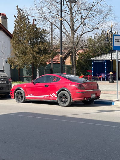 The scene depicts a static street view in Krivodol, Bulgaria, under clear skies with strong sunlight. The primary subject is a parked, modified red two-door sports coupe, possibly a Hyundai Tiburon/Coupe. The vehicle features black fender flares with visible rivets, black multi-spoke alloy wheels, and extensive white decals on its left side including "WRC" and a checkered flag pattern with "WORLD RALLY CHAMPIONSHIP" text. A black rear spoiler is affixed to the trunk lid, and the rear bumper incorporates a diffuser-like element with dual exhaust pipes. The Bulgarian license plate "BE HKH 5035" is mounted on the rear. A black Hyundai SUV with Bulgarian license plate "P 5293 CH" is partially visible parked directly in front of the red coupe.

The vehicles are parked alongside an asphalt road with a distinct blue painted curb separating it from a light-colored sidewalk. In the background, to the left, is a white or light-colored building with a terracotta tiled roof and a chimney. Evergreen trees with dense foliage are interspersed with bare deciduous trees, indicating a season of late winter or early spring. Two ornate black street lamp posts with gold accents are visible, one in the mid-ground and another to the right, adjacent to a blue and white bus stop sign depicting a bus pictogram. Further to the right, a small structure with a blue awning and walls appears to be an outdoor café or kiosk, featuring red and purple plastic chairs and tables, and stacked beverage crates. No individuals are visible in the scene.