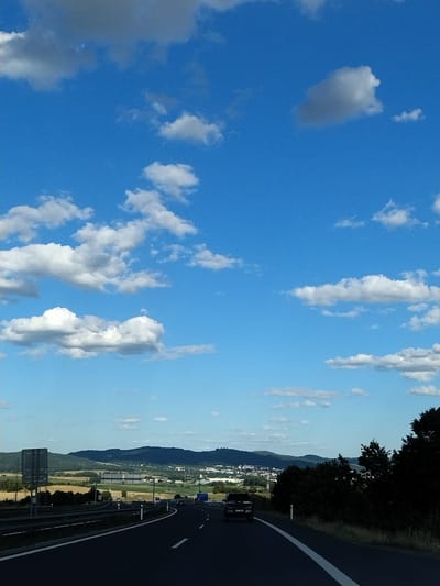 The image shows a view from a highway in Mladotice, Czechia, on a clear day. The sky is a vibrant blue with scattered white clouds. In the foreground, a dark asphalt road curves to the right, with white lane markings visible. On the left side of the road, there is a metal guardrail and a large gray traffic sign. A dark-colored SUV is driving on the road in the middle ground. Beyond the road, there are rolling hills covered in green trees and fields. In the distance, a town or city is visible nestled among the hills, with buildings and structures. The overall scene depicts a serene landscape with a focus on the highway and the natural environment.