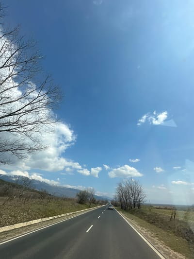 The scene depicts a rural road environment on a clear, sunny day, purportedly located in Osetenovo, Bulgaria. An asphalt two-lane road, marked with white dashed and solid lines, recedes into the distance, suggesting a travel perspective. A silver passenger vehicle is observed in the far distance, traveling in the same direction on the right lane. The roadside on both sides features dry, brown grass and sparse, leafless deciduous trees and shrubs, indicative of late winter or early spring. A concrete curb runs along the left edge of the road, bordering an elevated, sloping embankment. In the background, a range of mountains with snow-capped peaks is visible beneath a bright blue sky dotted with scattered white cumulus clouds. A faint crescent moon and a small, dark, distant object are discernible in the upper sky. Reflections, likely from a window, are present on the right side of the image. No individuals are visible within the frame.