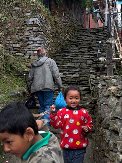 The image depicts a narrow, uphill stone staircase in the city of Ghandruk, Nepal, flanked by dry-stone walls. Three individuals are ascending these steps.
In the mid-ground, an adult male is seen from behind, wearing a grey hooded jacket, blue pants, and a patterned cap. He holds a blue plastic bag in his left hand and a closed black umbrella in his right hand.
In the foreground, to the right, a young girl with dark hair, approximately 5-7 years old, smiles directly at the viewer. She wears a red long-sleeved sweatshirt adorned with white and blue "Doraemon" cartoon characters and yellow bells, paired with dark pants. A small metal stud is visible in her left nostril. She holds a bundle of thin sticks in her right hand.
To the left of the girl, partially cropped by the image frame, is a young boy with dark hair, also approximately 5-7 years old. He wears a green-collared shirt beneath a camouflage-patterned hooded jacket. A small gold earring is visible in his left ear. He is looking downwards and to the left.
The stone steps are constructed from irregular flat grey stones, with sparse green vegetation growing between them and on the adjacent stone walls. In the upper background, beyond the adult, residential structures with orange-painted walls and traditional wooden balconies are visible, alongside a metal fence and wooden poles. A black pipe protrudes from the stone wall on the right side of the staircase.