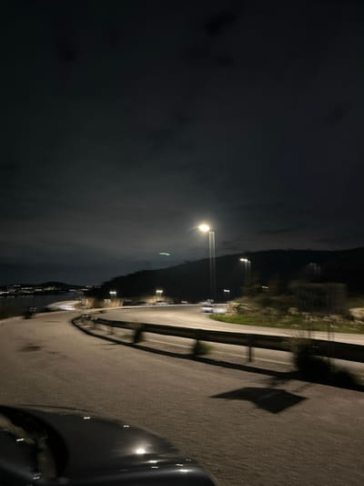 A night scene captured from the perspective of a vehicle shows a highway or road in Anatoliki Attiki, Greece. The image is dark and slightly blurry, suggesting movement or low light conditions.

In the foreground, part of a car's bodywork, possibly the hood or fender, is visible on the right, indicating the photo was taken from within or immediately beside a car. The midground features a multi-lane road or highway, illuminated by a series of streetlights that cast bright reflections on the apparently damp or reflective road surface. A metal guardrail runs along the side of the road, separating it from a lower, dark area, likely a body of water or a steep drop-off. Some light streaks are noticeable on the road, possibly from other moving vehicles or the effect of a long exposure.

The background consists of dark mountains or hills silhouetted against the cloudy night sky. Faint lights are scattered along the base of these distant landforms, hinting at distant towns or buildings. A large, bright orb, likely the moon, is prominently visible through the clouds in the upper left portion of the sky, acting as another significant light source. The overall weather appears to be cloudy, contributing to the hazy, dark atmosphere. There is no visible text in the image.