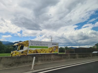 In St. Stefan ob Leoben, Austria, a yellow and white truck with "WIR KAUFEN IHR ALTSPEISEFETT" (We buy your used cooking oil) and "MÜNZER" branding is seen driving on a highway. The truck has a graphic of flowing oil on its side. The truck is traveling alongside a concrete barrier. In the background, there are trees, hills, and a power line tower. The sky is partly cloudy with patches of blue.