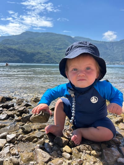 An infant is positioned in a low squat on a rocky shoreline, facing directly towards the camera. The infant has light-colored hair, blue eyes, and rosy cheeks, and is wearing a navy blue bucket hat, a two-toned blue long-sleeved rash guard featuring a white sailboat logo on the chest, and navy blue swim shorts. A grey beaded pacifier clip, with a grey animal-shaped attachment, is clipped to the rash guard and extends to a light brown pacifier resting near the infant's left foot. The infant's left hand is grasping a grey rock. The immediate foreground and midground consist of a wet, rocky beach and shallow, clear water. A large body of water, consistent with Lake Como, stretches behind the infant. In the far left background, a person's head and shoulders are visible above the water line. Across the lake, steep, forested mountains rise prominently. The sky above is blue with scattered white clouds. The scene is illuminated by direct sunlight, indicating a clear day in Abbadia Lariana, Italy.