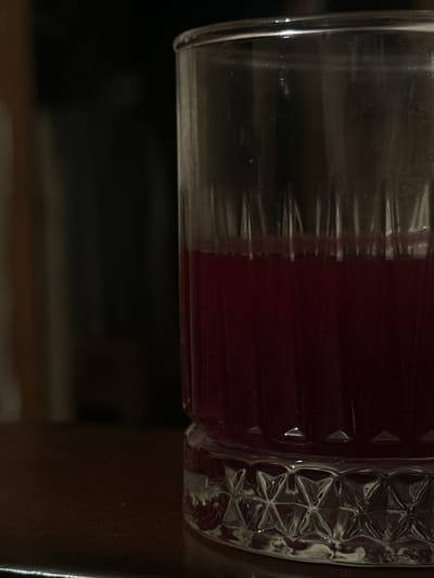 The image displays a close-up of a clear glass containing a dark red liquid, positioned horizontally. No people are visible in the frame.

The glass features two distinct textures: one side is smooth and transparent, showcasing reflections and refractions through its surface, while the other side is adorned with an intricate, repeating pattern of facets, possibly star-shaped or geometric designs, which sparkle under the light. The dark red liquid, possibly juice or wine, fills roughly half of the visible portion of the glass.

The background is dark and blurry, suggesting an indoor setting under low light conditions, such as evening or night. There are no specific location cues within the image to indicate Agarak, Armenia, beyond the provided context. No activity is taking place, and no text is visible on the glass or in the background.