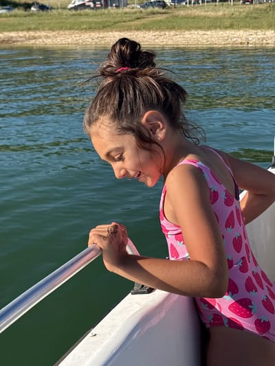 A young girl with dark hair pulled back in a bun is on a boat. She is wearing a pink swimsuit with a strawberry pattern. The boat is on the water, and a sandy shore with green vegetation is visible in the background. The sun is shining, casting a shadow on the girl's legs. The girl appears happy, with a smile on her face. The water is a deep blue-green. The scene suggests a sunny day, likely in the warmer months, possibly a vacation or day trip.