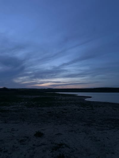 A vertical, long shot shows a twilight scene, likely in Trastikovo, Bulgaria, showcasing an overcast sky in gradations of blue and gray. The sky transitions to a slight orange tint along the horizon where it meets a body of water to the right. Adjacent to the water is a dark, indistinct shoreline or bank, which could be vegetation or rocky terrain. Details of the land are obscured due to the low light conditions. No people, specific objects, actions, or interactions are visible. The environment dominates the frame, depicting a natural setting at dusk or dawn.