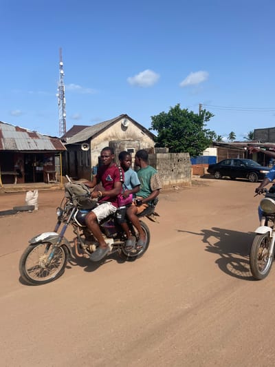 Three people are riding on a motorcycle on a dirt road. They are near a building with a corrugated metal roof and a communications tower. A car is parked along the road. The location appears to be in Ipota, Nigeria.