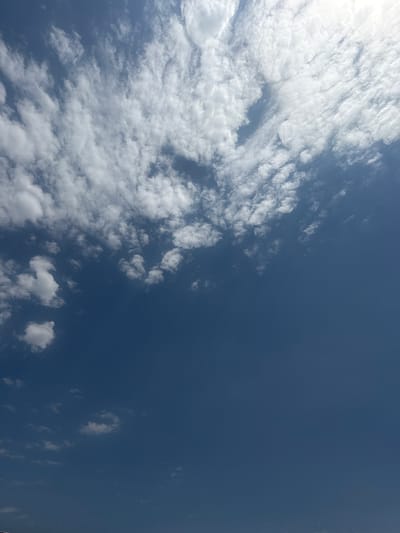 The image is a close-up, low-angle shot of a blue sky with scattered white clouds. The clouds are wispy and appear to be high in the atmosphere. The lower portion of the image is a solid expanse of blue sky, with a few smaller clouds visible near the bottom. There are no people, structures, or geographical features visible in the image. The location in Kashkarlik, Uzbekistan, is not discernible from the image content.