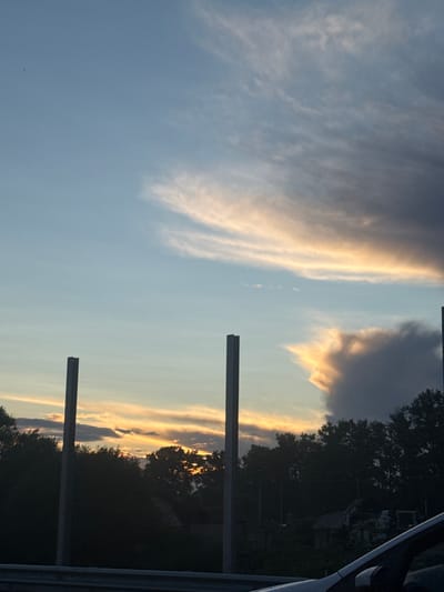 The image captures a twilight sky over Krutitsy, Russia, likely from the perspective of a vehicle. The sky is a canvas of soft blues and oranges, with wispy clouds streaked with sunlight, hinting at the setting sun. Two tall, dark, rectangular pillars, possibly part of a fence or structure, stand in the foreground, bisecting the view. Beyond them, a dense line of dark trees silhouettes the horizon. The overall atmosphere is serene, with the natural beauty of the sky contrasting with the man-made structures. There are no people visible in the frame.