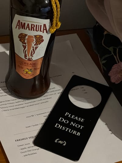 A still-life shot depicts a bottle of Amarula Cream liqueur positioned beside a menu and a "Please Do Not Disturb" door hanger. The Amarula bottle, sealed with a gold cord, is partially obscured, focusing on its label, which features the Amarula brand name, an elephant graphic, and details like "CREAM WITH MARULA SPIRIT" and "750 ml". The menu underneath displays breakfast options, including "Summer Frittata", "Savoury mince omelette", "Creamy scrambled eggs", and "Freshly Squeezed" juice. The black door hanger with "PLEASE DO NOT DISTURB" in white capital letters lies on the menu. Based on the menu items, the scene could potentially be set at a hotel or guesthouse in Overstrand Local Municipality, South Africa.