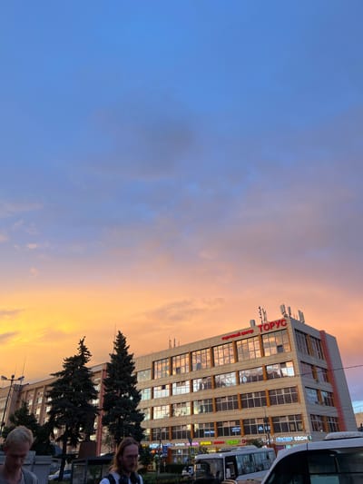 The image captures a street scene in Лесные Поляны, Russia, during sunset. The sky is painted with vibrant hues of blue, orange, and yellow, indicating a picturesque end to the day. A modern, multi-story building with numerous windows dominates the right side of the frame. The windows reflect the colorful sky, adding to the atmospheric effect. A red sign on the building reads "ТОРУС," and below it, "Торговый Центр" (Trade Center). Several other signs with text, some in Cyrillic, are visible on the building's lower levels, including what appears to be "OZON."
In the foreground, two people are partially visible, looking towards the right of the frame, seemingly observing something or someone out of view. One person is wearing a black and white checkered garment, and the other has lighter hair. Dark green coniferous trees stand between the building and the street, adding a natural element to the urban landscape. The overall mood is serene, illuminated by the warm glow of the setting sun. The presence of a bus on the right suggests it might be a public transport hub or a busy street. The time of day is clearly sunset, and the weather appears clear with scattered clouds contributing to the dramatic sky.