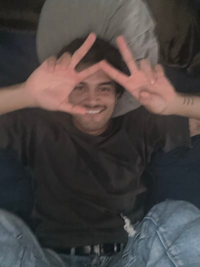 The image depicts a young man, likely in his late teens or early twenties, making the peace sign with both hands in front of his face. He is smiling and wearing a black t-shirt and blue jeans. The man appears to be lying on a bed or couch with a gray pillow behind him. The location is a bedroom in Celaya, Mexico, based on the provided coordinates. The image is blurry and dimly lit, and the subject's face is obscured by his hands. The background is partially obscured by the subject's body. The subject's left leg is visible in the image, as well as a portion of his right arm. There appears to be a tattoo on the man's right wrist.  There are no other people or objects visible in the image. The man's posture suggests he is relaxed and comfortable.