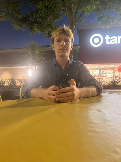 An individual, identified as a young male with light-colored hair and a dark long-sleeved shirt, is seated at a mustard-yellow outdoor table, facing the camera directly. He holds a dark-colored smartphone with both hands, resting on the table surface. A silver-colored bracelet is visible on his left wrist. The background, illuminated at dusk or night, features a large tree with green foliage directly above him. To his right, a Target retail store is visible, identified by its illuminated white "Target" logo and red facade, with brightly lit automatic sliding doors marking the entrance. To his left, another commercial building with a dark facade and warm-toned exterior lighting is present. Various landscape plantings, including a large spiky-leaved plant and smaller shrubs, are situated along the base of the buildings. The scene is located outdoors in a commercial area of Rolling Hills Estates, United States, likely an exterior dining or seating space.