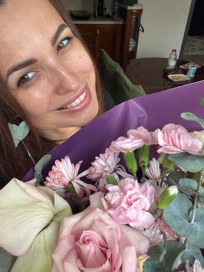 A close-up, waist-up shot captures a woman with long, brown hair and light skin smiling while holding a bouquet of pink and white flowers. She is positioned in the foreground, with a blurred background of a room. The woman's head is tilted to her left, and her gaze is directed at the camera. She is wearing dark clothing. The bouquet, wrapped in purple paper, features roses, carnations, and eucalyptus leaves. Behind her, a green-backed chair is visible, as is a portion of a wooden table with a water bottle, snacks, and a book. The scene appears to be indoors, in what seems to be a residential setting.