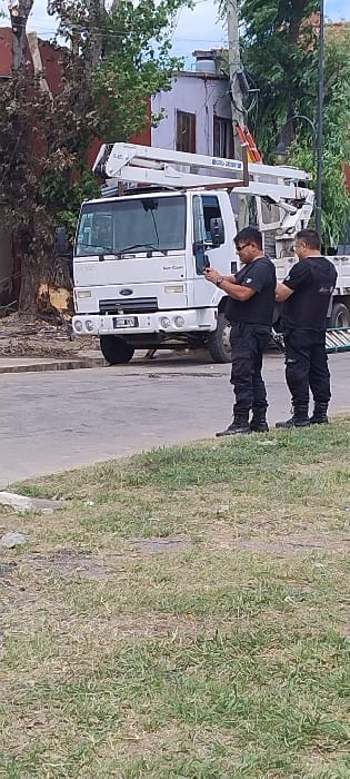 The image depicts an outdoor scene in what appears to be a residential or semi-urban area, potentially San Fernando, Argentina. Two men, dressed in dark uniforms consisting of black shirts, black pants, and black boots, are standing near a white utility truck equipped with a boom lift.

The utility truck, a Ford "Super Cargo" model as indicated by the visible branding, is parked on what seems to be a paved roadside. Its boom lift is retracted and positioned over the front of the vehicle. One of the men is positioned closer to the truck, looking down at something in his hands, possibly a device or paperwork. The second man stands slightly behind and to the right, observing the scene or the truck. Their attire and the presence of the boom lift truck suggest they are utility workers or technicians, likely involved in maintenance, repair, or installation tasks related to overhead lines or structures.

In the background, behind the truck and the men, are residential buildings or houses with visible windows and roofs. Utility poles with wires are also visible, hinting at the nature of the work. Lush green trees and vegetation frame the left side of the scene. The foreground consists of a paved surface adjacent to a grassy area with varying shades of green and brown. The natural lighting indicates it is daytime with clear weather. No strong emotions are discernible from the individuals, who appear focused on their activity.