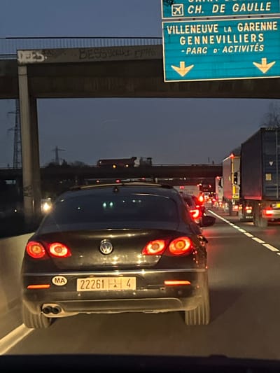 A black Volkswagen is stuck in traffic in France. The car has a Moroccan license plate that reads "22261 A 4". A blue overhead road sign indicates directions for Villeneuve La Garenne, Gennevilliers, and Parc D'activités. The sign also points to Ch. de Gaulle. Graffiti is visible on a concrete barrier near the road. The traffic ahead is heavy and congested.